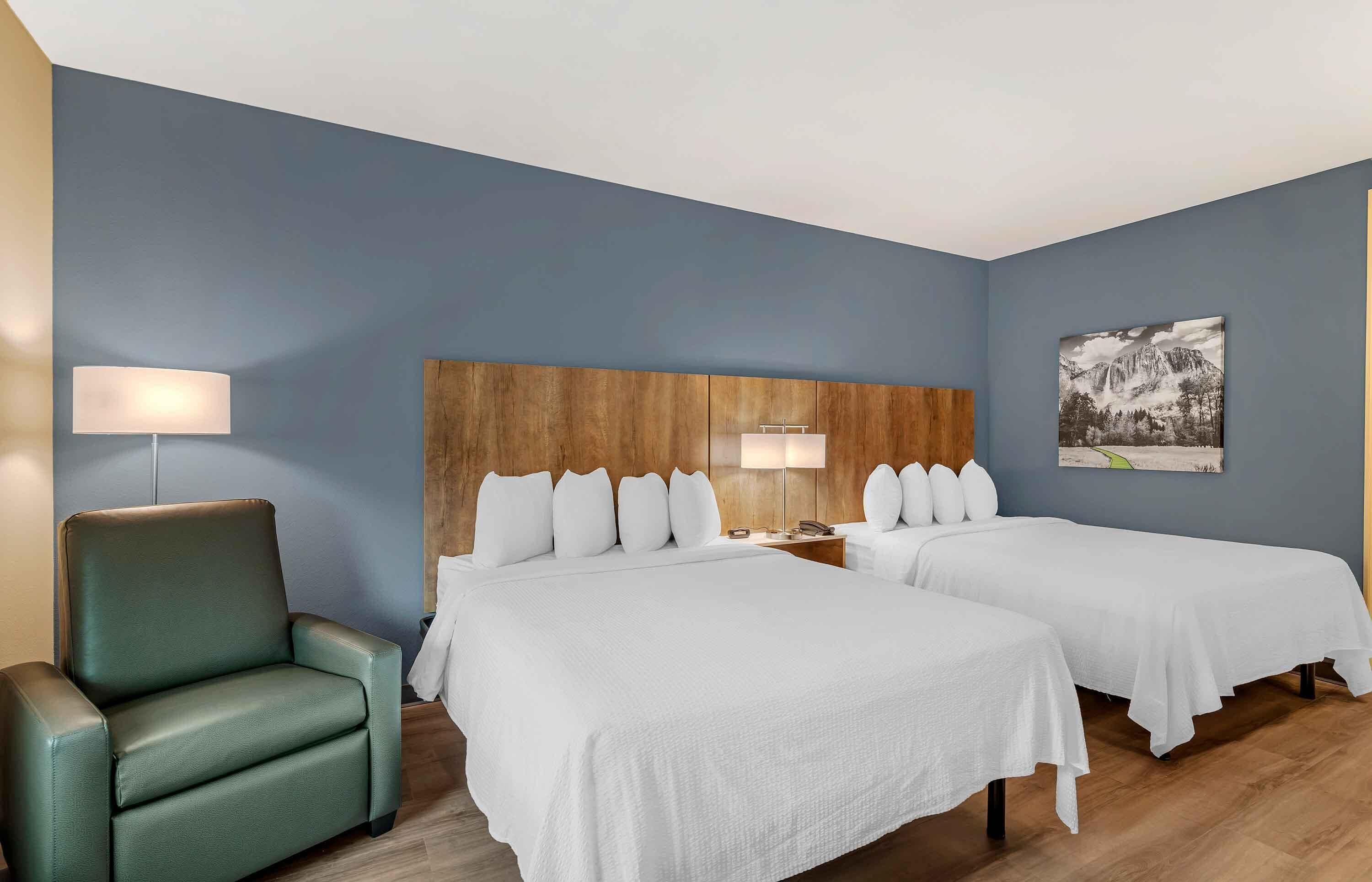 Habitación de hotel con dos camas, cabecero de madera, sillón verde y un cuadro con paisaje de montaña en la pared.