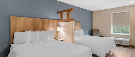 Habitación de hotel con dos camas, cabecero de madera, mesita de noche, lámpara, silla, ventana y radiador.