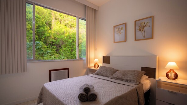 A neatly made bed with rolled towels, a large window with curtains, two bedside lamps, and two framed pictures on the wall. - SUH Copacabana 63 Peace Beach (Rio de Janeiro)