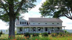 A two-story white house with a porch, surrounded by greenery and flowers. - Hoppy Hill Farm House (Dover Plains)
