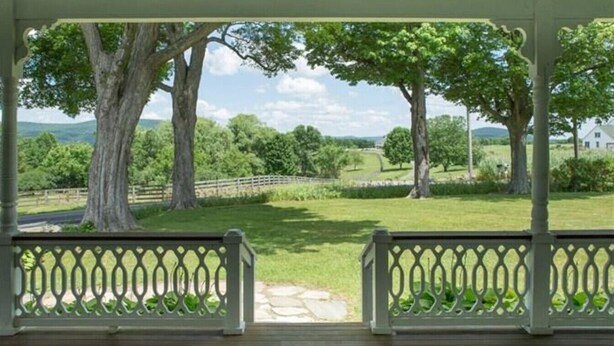 A covered outdoor area with a wooden deck and a view of a grassy landscape with trees and a distant white building. - Hoppy Hill Farm House (Dover Plains)
