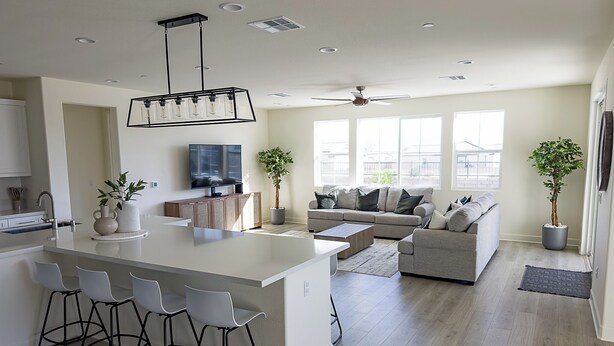 A modern kitchen with a central island, bar stools, a dining area, a living room with a sofa and a TV, and two potted plants. - The Palms Hideaway - Desert Luxury (La Quinta)