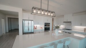 A modern kitchen with white cabinetry, a large island with bar stools, stainless steel appliances, and a pendant light fixture. - The Palms Hideaway - Desert Luxury (La Quinta)