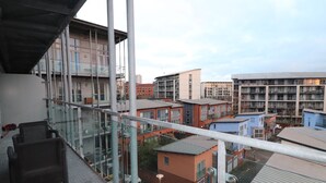 A modern building with a glass balcony, a wicker chair, and a view of surrounding buildings. - Exceptional Two-Bedroom Apartment in the City Center. (Birmingham)