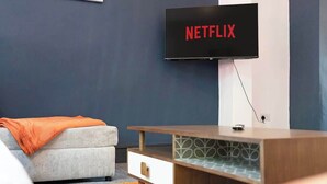 A modern living room with a flat-screen TV mounted on the wall displaying 'NETFLIX,' a grey sofa with an orange throw blanket, and a wooden side table with a drawer.