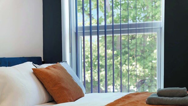 A bed with white and orange pillows, a brown blanket, and a view of trees through a window with vertical blinds.