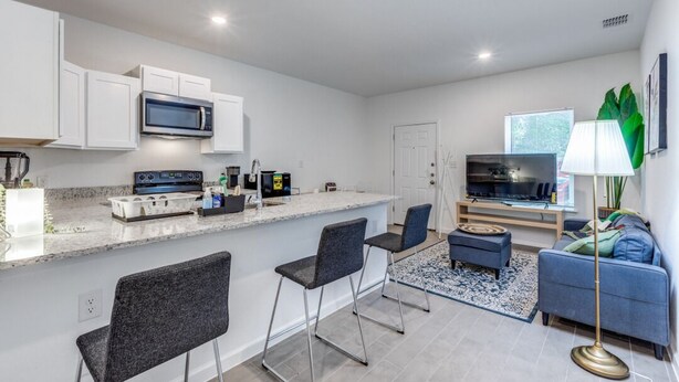 A modern kitchen with white cabinets, a microwave, and a built-in oven. There are two bar stools and a small dining table. A sofa and a floor lamp are in the living area.