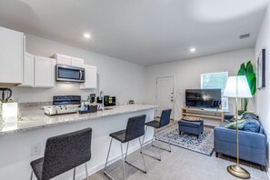 A modern kitchen with white cabinets, a microwave, and a built-in oven. There are two bar stools and a small dining table. A sofa and a floor lamp are in the living area. - Casa Nueva 3 Habitaciones, Renta por cuarto o toda, a solo 7 min de downtown. (San Antonio)