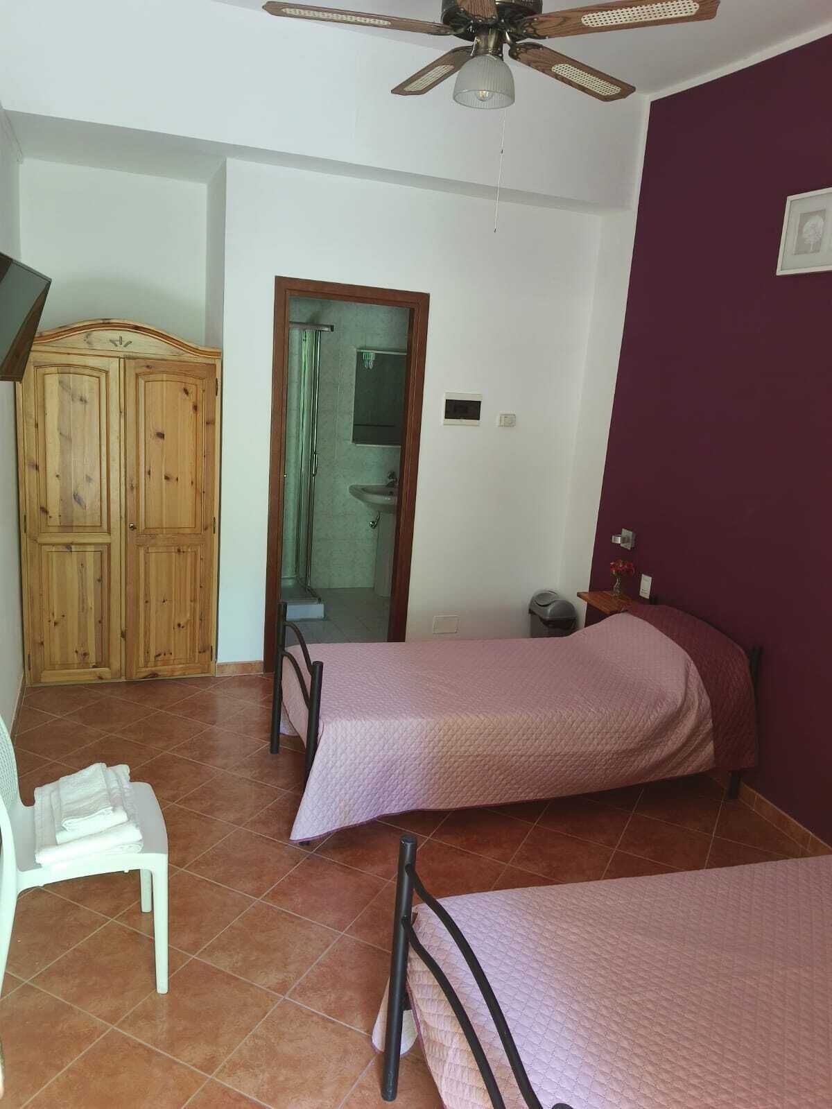 Un dormitorio con dos camas, un armario de madera, un baño con puerta de vidrio y un ventilador de techo.