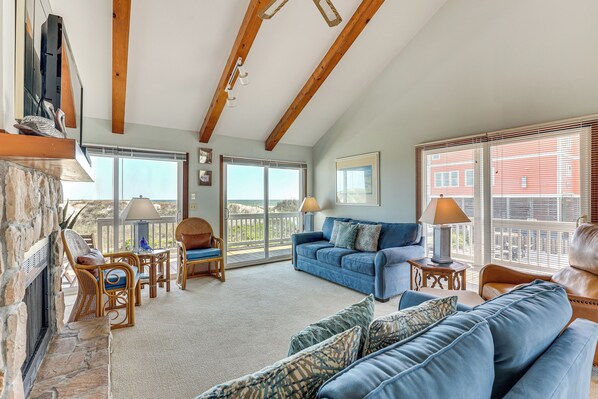 A spacious living room with a blue sofa, wicker chairs, a fireplace, and large windows offering a view of the outside. - Deck & View: Oceanfront Oasis in Oak Island! (Oak Island)