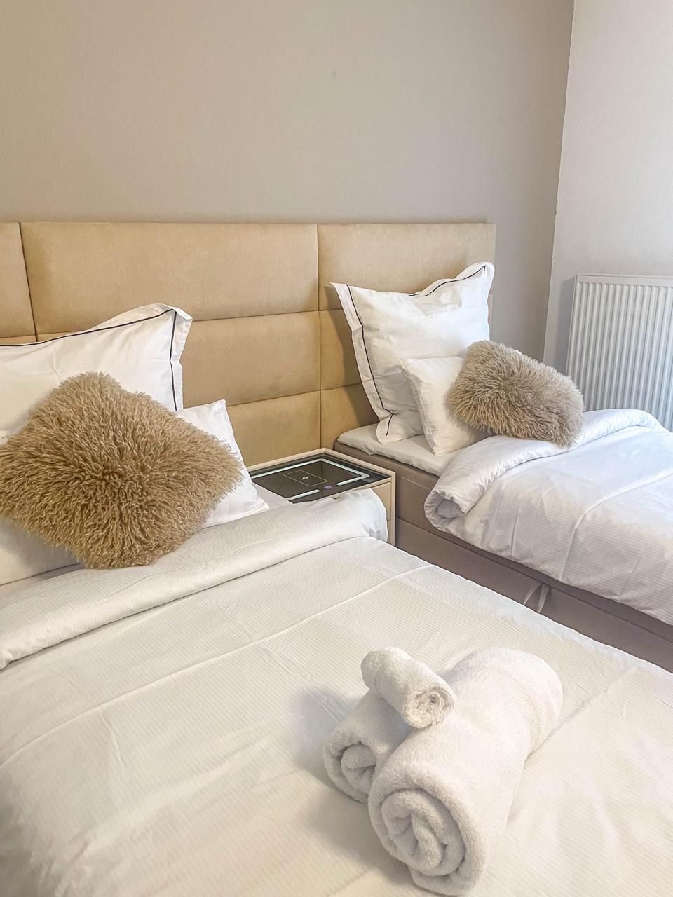 A hotel room with two beds, each with white linens and fluffy pillows. There is a bedside table with a power outlet and a small lamp.