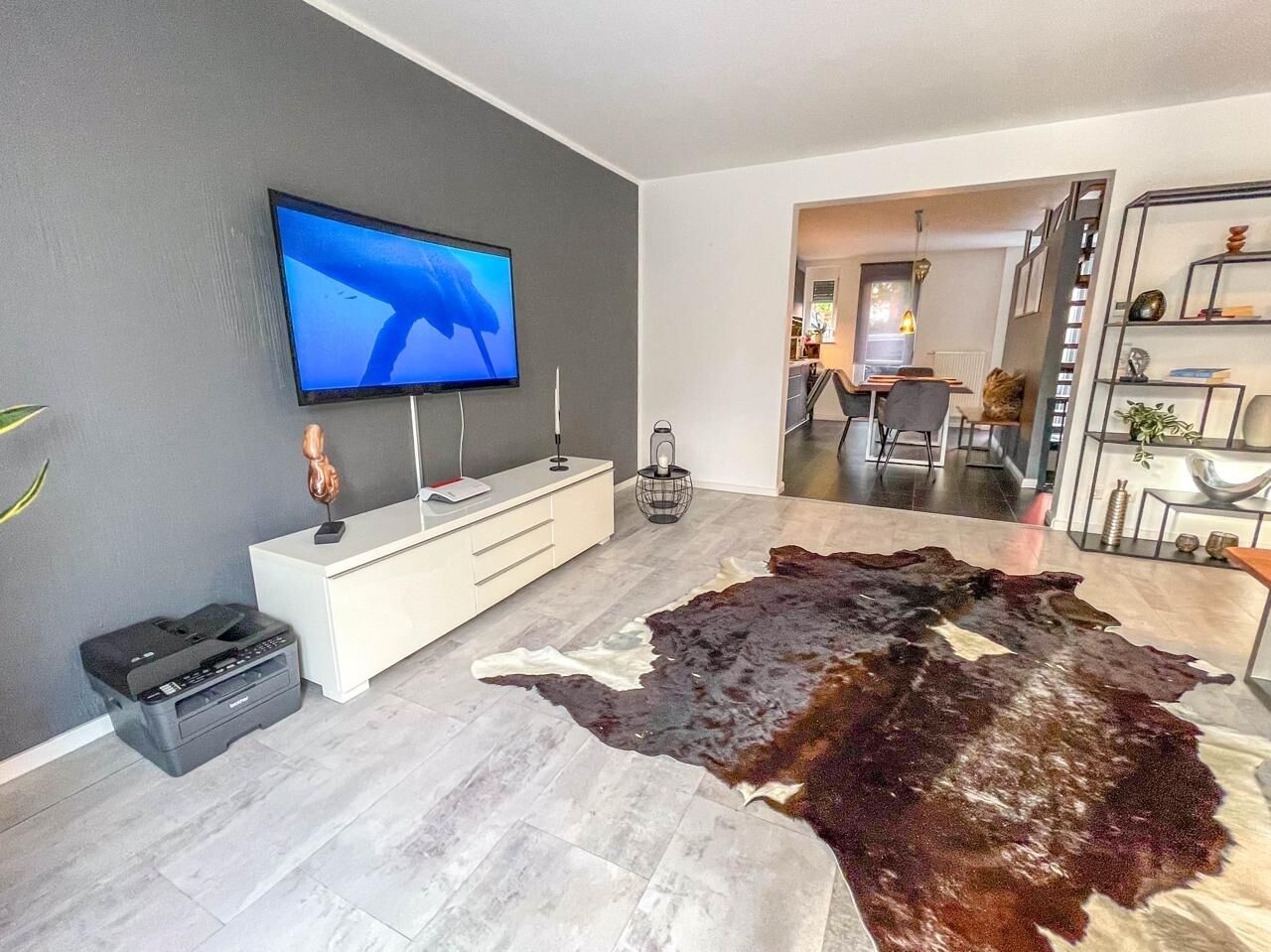 Ein modernes Wohnzimmer mit einem großen Flachbildfernseher an der Wand, einer weißen Unterhaltungsanlage, einem Lederteppich und einer Treppe, die in einen anderen Raum führt.