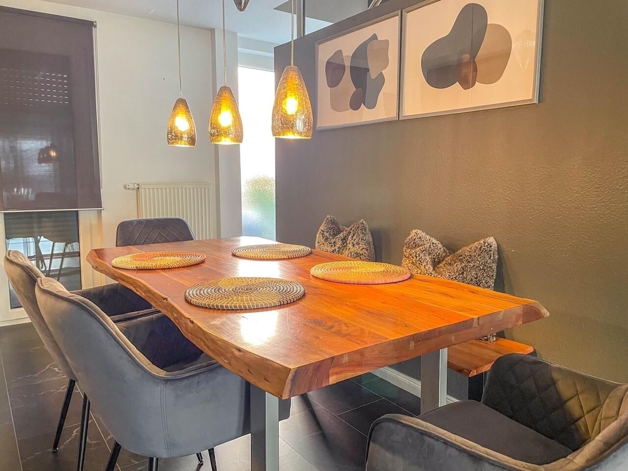 Un espace repas avec une table en bois, des chaises noires à coussin, des sets de table en fibres tressées et des luminaires suspendus.