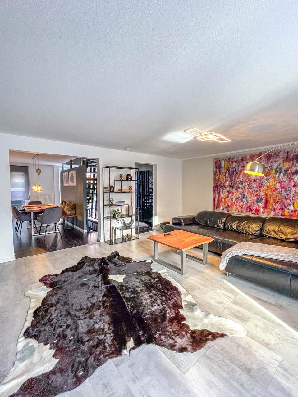 A modern living room with a leather sofa, a coffee table, and a large rug. There is a dining area in the background with a table and chairs.