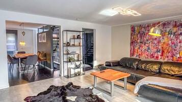 A modern living room with a leather sofa, a coffee table, and a large rug. There is a dining area in the background with a table and chairs.
