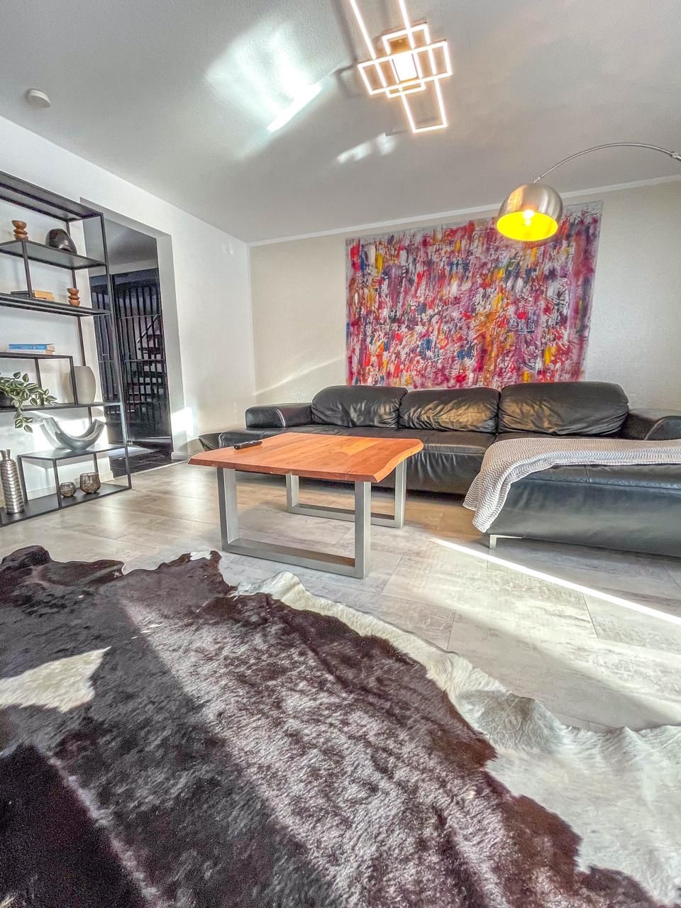 Een moderne woonkamer met een leren bank, een houten salontafel, een kleden van runderleer en een kleurrijk abstract schilderij aan de muur.