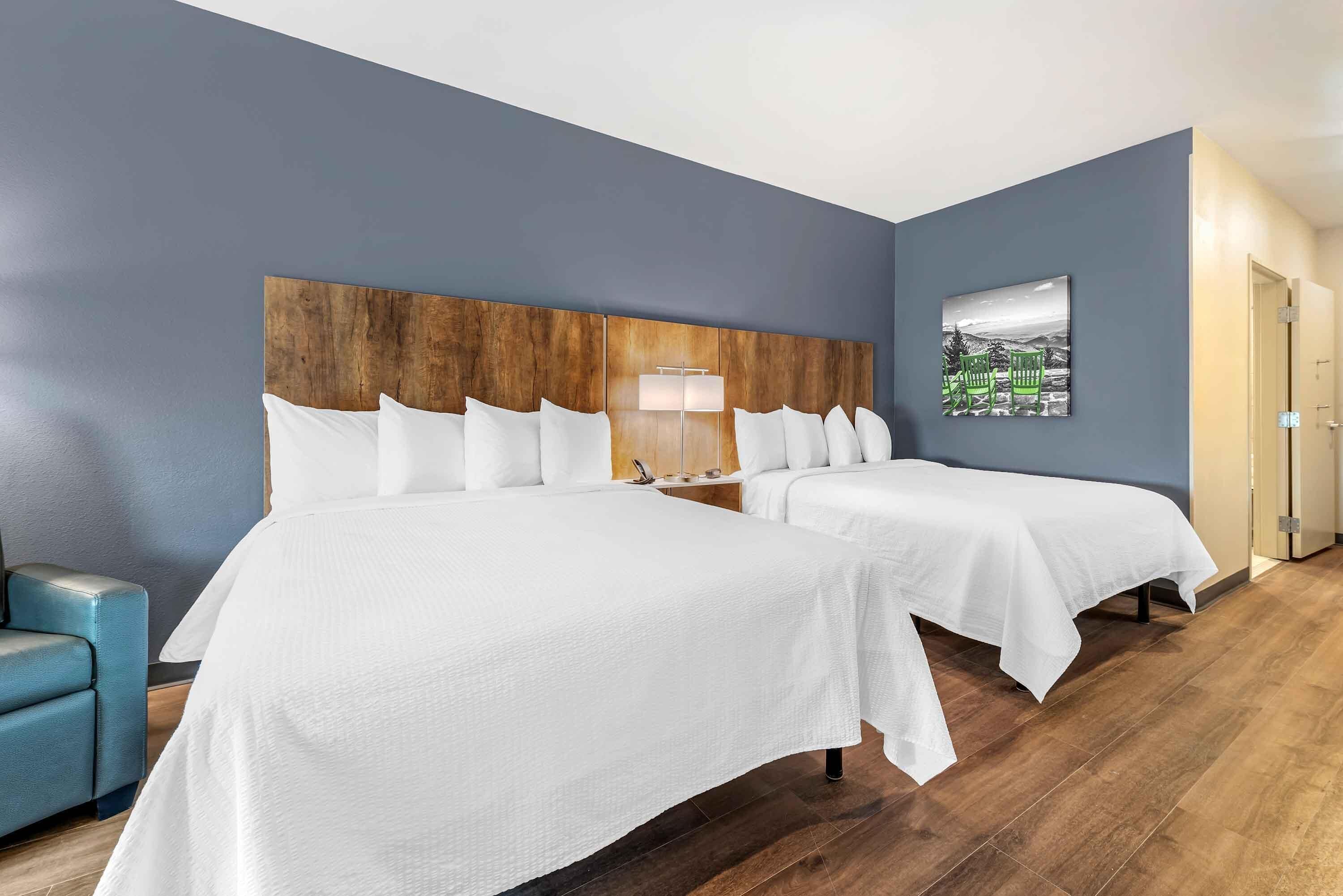 A hotel room with two beds, a wooden headboard, a blue wall, a green bench, and a painting.