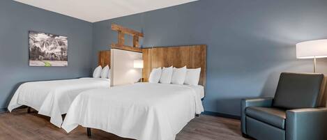 Habitación de hotel con dos camas, cabecero de madera, un sillón azul y una lámpara fijada a la pared.