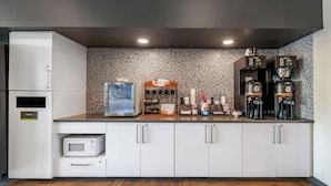 Una cocina moderna con gabinetes blancos, microondas, un cubo de basura, una estación de café con dos cafeteras y un salpicadero decorativo.