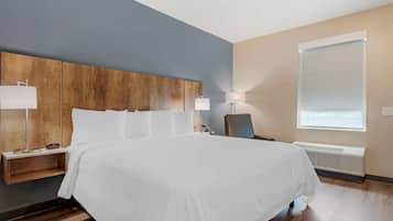 A hotel room with a large bed, wooden headboard, bedside tables with lamps, a chair, and a window with blinds.