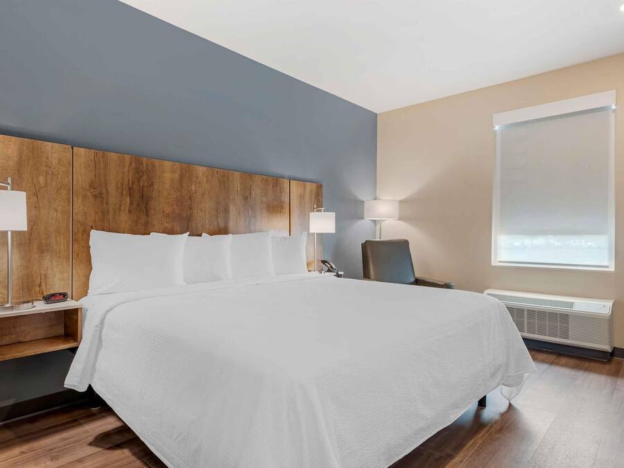 A hotel room with a large bed, wooden headboard, bedside tables with lamps, a chair, and a window with blinds.