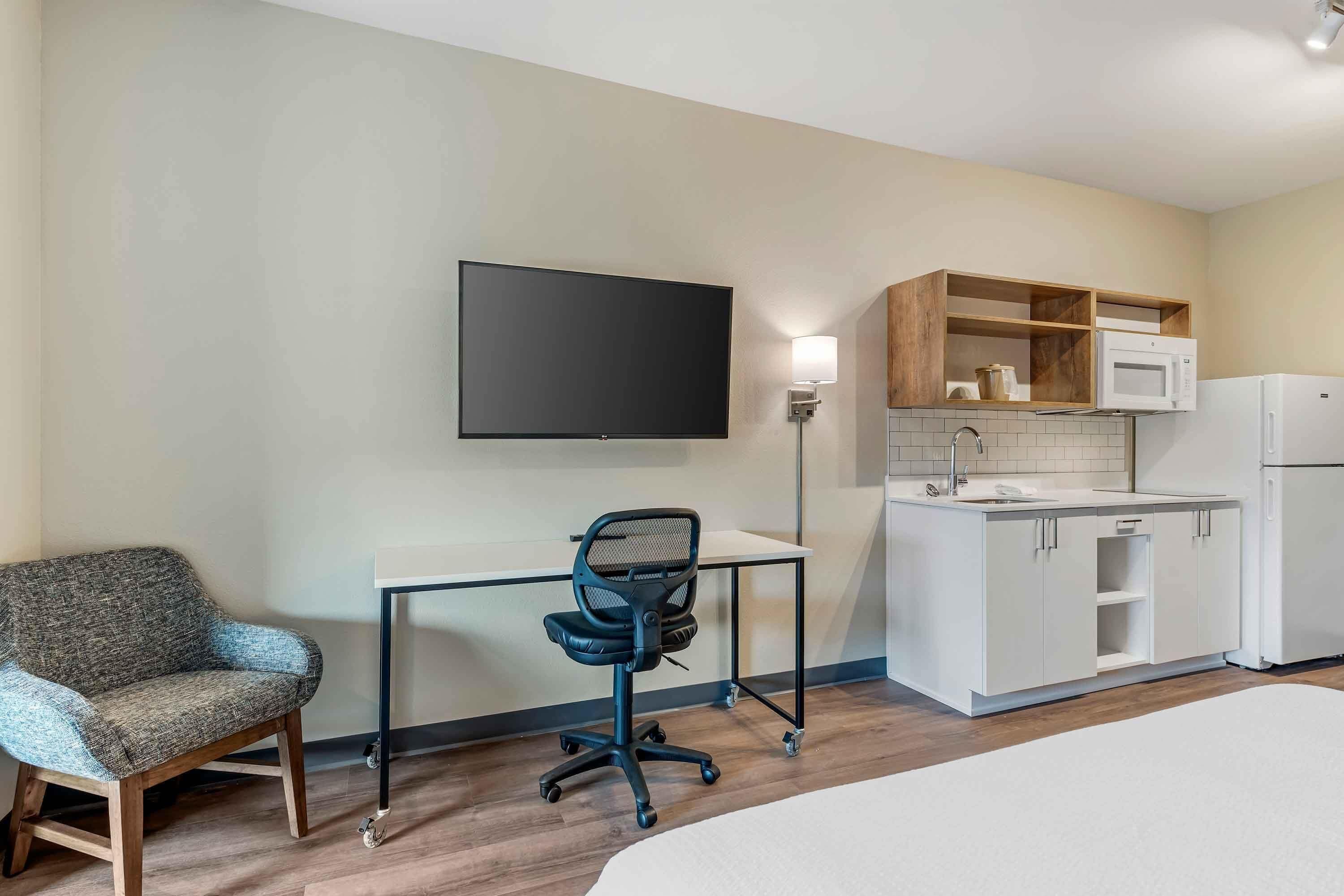 Hotelkamer met een bed, een bureau met stoel, een flatscreen-tv, een kitchenette met een magnetron en koelkast, en een houten plank boven het aanrecht.