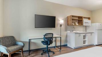 Hotelkamer met een bed, een bureau met stoel, een flatscreen-tv, een kitchenette met een magnetron en koelkast, en een houten plank boven het aanrecht.