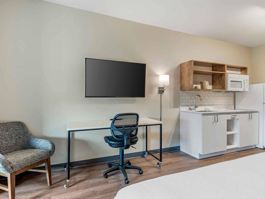 Hotelkamer met een bed, een bureau met stoel, een flatscreen-tv, een kitchenette met een magnetron en koelkast, en een houten plank boven het aanrecht.