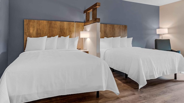 Habitación de hotel con dos camas, cabeceros de madera, ropa de cama blanca, lámparas de noche y una silla.