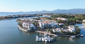 A coastal marina with modern buildings, boats docked, and a clear view of the sea and mountains in the background.