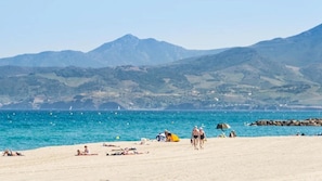 A beach with people sunbathing and walking, a rocky shoreline, and mountains in the background.