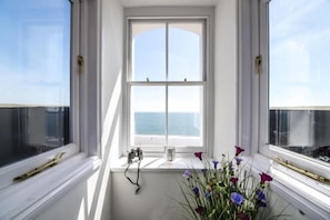 A room with a large window overlooking the sea, a small window sill with a vase of flowers, and a wall-mounted camera.
