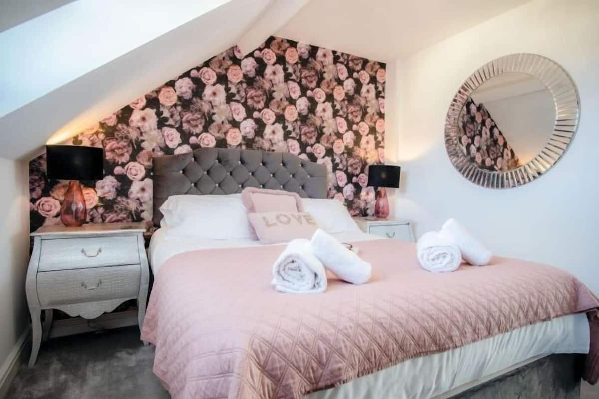 A bedroom with a floral wallpaper, a tufted headboard, a round mirror, and a bed with pink bedding and rolled towels.