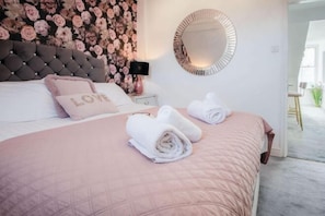 A neatly made bed with rolled towels, a tufted headboard, a floral wallpaper, and a round mirror on the wall.