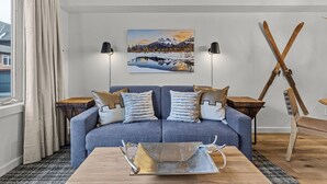 A modern living room with a blue sofa, two side tables, a wooden coffee table, a painting of a snowy landscape, and a pair of skis leaning against the wall. - Alpine Loft Penthouse [Outdoor Heated Pool & x3 Hot Tubs] (Canmore)