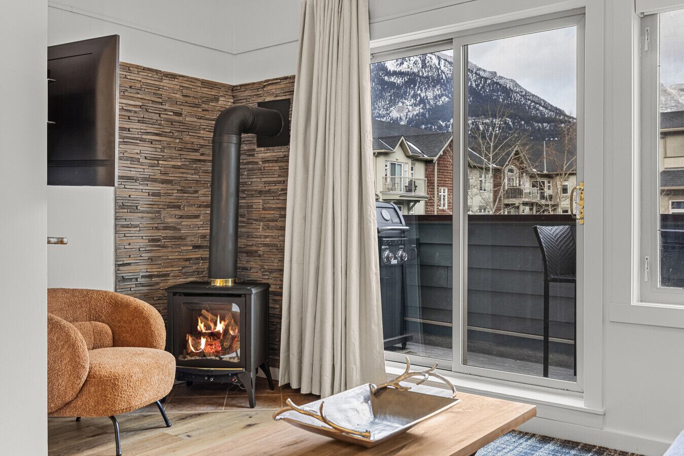 A cozy living room with a wood-burning stove, a comfortable armchair, and a view of a snowy mountain through a large window.