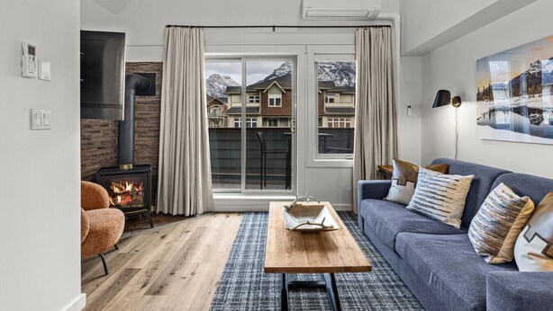 A modern living room with a fireplace, a sofa, and a view of a mountain landscape through a large window. - Alpine Loft Penthouse [Outdoor Heated Pool & x3 Hot Tubs] (Canmore)