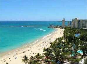 A beach with clear turquoise waters, a sandy shore, and palm trees. There are high-rise buildings in the background. - Apartment with Sea View in Isla Verde (Carolina)