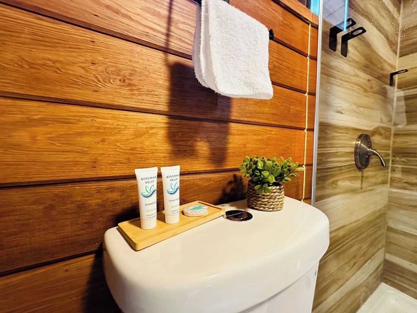A bathroom with wooden panel walls, a white toilet, a small potted plant, and two bottles of Bergman Kelly shampoo and conditioner on the toilet tank.