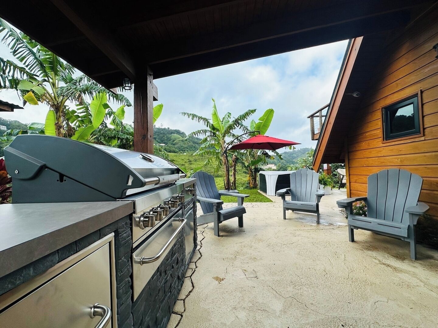 An outdoor patio with a grill, two Adirondack chairs, and a red umbrella.
