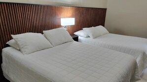 Une chambre d’hôtel avec deux lits, chacun doté de draps et d’oreillers blancs. On y trouve une tête de lit en bois et une table de chevet avec une lampe.