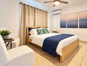 A bedroom with a bed, a ceiling fan, a nightstand, a chair, a potted plant, and a wall-mounted air conditioner.
