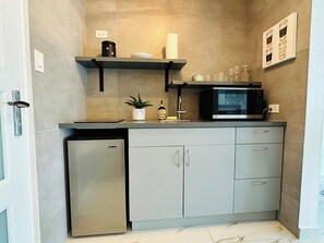 Una cucina compatta con lavastoviglie integrata, forno a microonde e un piccolo frigorifero. Sono presenti mobili con maniglie e uno scaffale sopra il piano di lavoro.