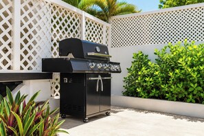 A portable grill with wheels, set against a white lattice fence, with lush green foliage in the background. - CasaPlaya Gamil Rene (Barceloneta)