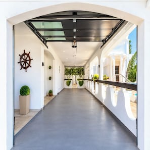 A corridor with a glass ceiling, a ship's wheel decoration, potted plants, and a view of a pool area. - CasaPlaya Gamil Rene (Barceloneta)