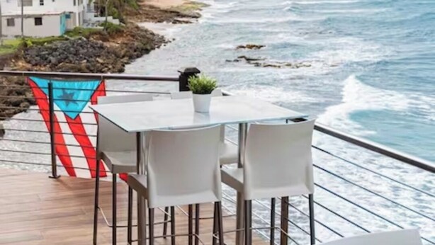 A deck with a table and chairs overlooking a beach with waves, a flag, and buildings in the background.