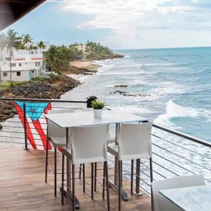 A deck with a table and chairs overlooking a beach with waves, a flag, and buildings in the background. - Luxury Ocean Villa Steps from the Beach (Dorado)