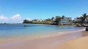 A beach with clear water, a sandy shore, and buildings in the background.