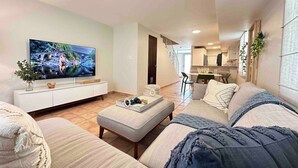 A modern living room with a flat-screen TV, a sofa, a coffee table, and a dining area in the background.