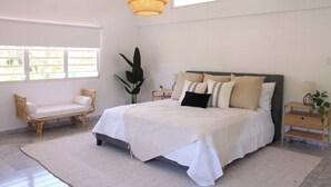 A bedroom with a large bed, a bedside table, a chair, a hanging light fixture, and a window with blinds. - Estancia Nueva Colonial (Manati)